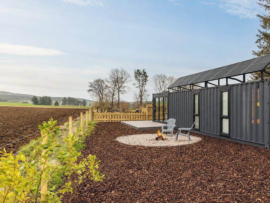 Dark grey container home beside a fire pit at the edge of a ploughed field